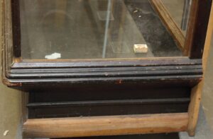 Close-up of an antique glass-front showcase with an oak frame and black painted beveled base, showing worn edges and a small support block inside.