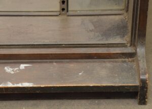 Close-up of a dark wood tone tabletop showcase with a mirror back and worn surface, showing the lower front edge and built-in shelf tracks.