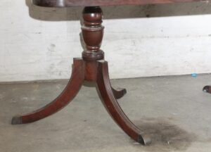 Close-up of a Regency Revival dining table base with an urn-shaped center pillar and three curved saber legs in a dark wood tone.