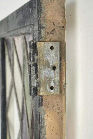 Close-up of a steel casement window hinge with worn galvanized surface, black-painted frame edge, and part of the leaded stained glass panel visible beside it.