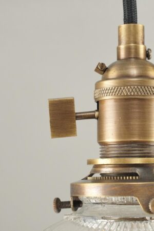 Close-up of an industrial pendant light with an antique brass fitter, black cord, and clear Holophane-style glass shade.