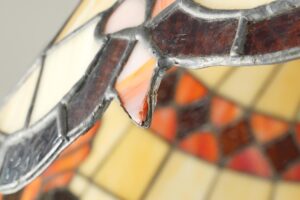 Close-up of a Tiffany style pendant light shade showing stained glass segments in cream, amber, and orange with dark leading and a small damaged section.