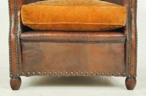 Vintage brown leather club chair with a plush orange seat cushion, brass stud trim, and turned wooden legs.