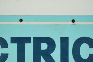 Close-up of a vintage wooden commercial sign painted blue and white, showing the upper holes and the large dark lettering “TRI.”