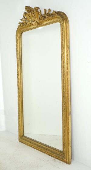 Tall giltwood mirror with rounded upper corners, an ornate crest, and visible age spots on the glass.
