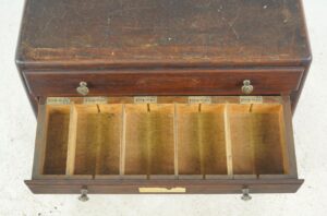 Open drawer of a vintage industrial display cabinet made of dark wood, showing six divided compartments with small handwritten labels and brass pulls.