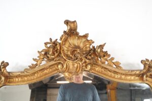 Close-up of an antique Louis XV Rococo wall mirror, showing the ornate gilded crest with shell and curling foliage details and some visible wear on the frame.