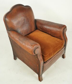 Art Deco club chair with a mustache-shaped back, rounded arms, and a velvet seat cushion, shown in three-quarter view with visible wear and studded trim.