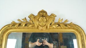 Antique French Louis Philippe giltwood mirror with rounded upper corners and a straight bottom edge, shown in close-up with an ornate crest and visible age spots on the glass.