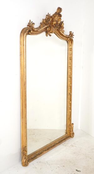 Antique Louis XV Rococo wall mirror with a gilt frame and ornate shell and foliage crest, shown at an angle with visible age wear and some missing decorative details.
