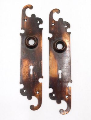 Pair of brass door back plates with a traditional swirl shape and keyhole cutouts, shown as a matching pair with black and copper finish wear.
