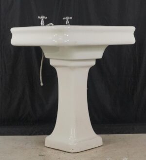 White Art Deco pedestal sink with chrome hardware, shown front-on with a wide basin, tapered base, and visible surface wear.