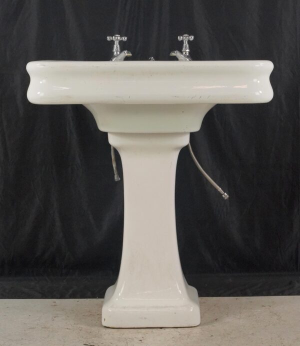 White porcelain pedestal sink with chrome cross-handle faucets and exposed supply lines, shown from the front with visible surface wear and staining.