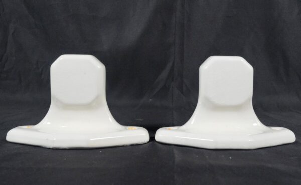 Pair of white ceramic towel bar brackets shown side by side, each with an octagonal top and wide base, with visible screw holes and slight surface wear.