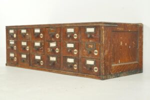 Antique tabletop card catalog cabinet with eighteen small drawers, brass label holders, and round knobs, shown in a three-quarter view with the side panel visible.