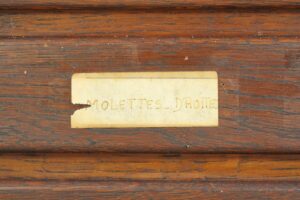 Close-up of a vintage industrial storage cabinet drawer label on dark wood, showing a worn cream paper tag with handwritten text.