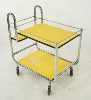 1950s mid century modern rolling bar cart with chrome frame, two yellow Formica shelves trimmed in black, and four caster wheels shown at an angle.