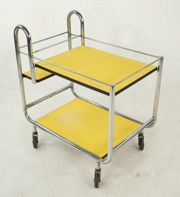 1950s mid century modern rolling bar cart with chrome frame, two yellow Formica shelves trimmed in black, and four caster wheels shown at an angle.