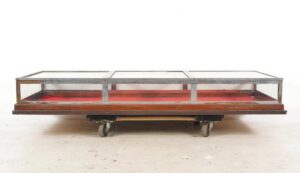 Mid century vitrine display case with a clear glass top, dark wood base, and red felt interior, shown in a front view with surface wear and a broken pane visible.