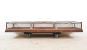 Mid century vitrine display case with a lift-up top, clear glass panels, and red felt interior, shown in a wide front view on casters with visible surface wear.