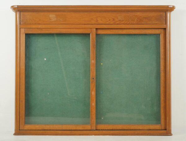 Wall-mounted display case with an oak frame, two glass sliding doors, and a green cork board insert.