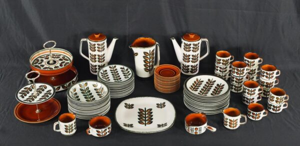 A 1960s BOCH Rambouillet dishware set is arranged with stacked plates, bowls, cups, pitchers, serving pieces, and two tiered stands, all featuring a leaf pattern in black, brown, and white.