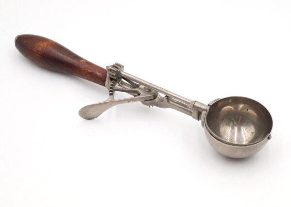 Antique ice cream scoop with a dark wood handle and nickel-plated metal bowl, shown in profile with the thumb lever and stamped shaft visible.
