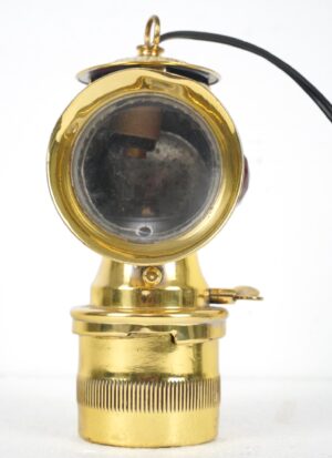 Antique converted carbide bicycle lamp with a polished brass body, round clear lens, and attached power cord, shown from the front.