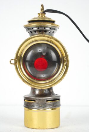 Antique converted carbide bicycle lamp with a brass and steel body, a clear front lens with a red center, and a cord attached at the top.