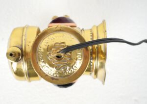 Antique carbide bicycle lamp in polished brass with a black cord, shown from the top with the red lens visible and signs of surface wear.