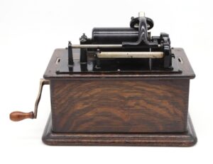 Antique phonograph with a dark wood base, black and brushed steel mechanism, and a hand crank on the left side.