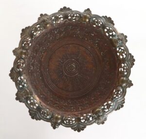 Victorian brass plant stand top with a scalloped, embossed edge and circular floral center detail, shown from above with visible tarnish and age-related wear.