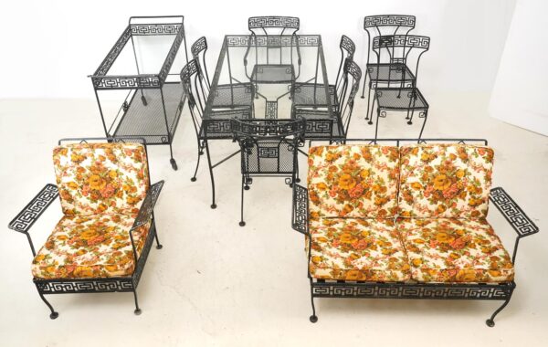 Twelve-piece mid-century patio furniture set with black wrought iron Greek key frames, including a glass-top table, chairs, a serving cart, and cushioned seating with floral cushions visible on the front pieces.