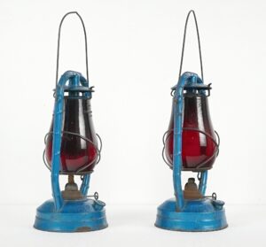 Pair of vintage kerosene lanterns with blue painted steel frames and ruby red glass globes, shown side by side with arched wire handles and visible wear.