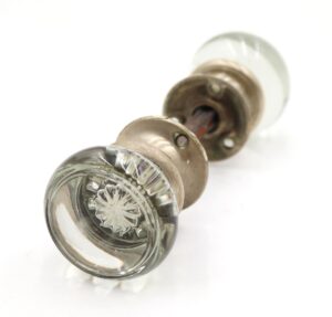 Clear glass door knob set with brushed nickel rosettes shown in close-up, featuring one knob in the foreground and the matching knob behind it on a spindle.