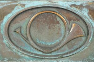 Antique bronze window panel with a verdigris finish, featuring a central post horn relief and heavy surface wear.