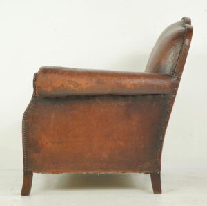 Side view of a vintage Art Deco club chair with a rounded armrest, studded trim, and visible wear on the brown leather upholstery and wooden legs.