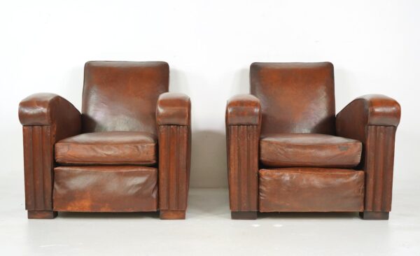 Pair of vintage Art Deco leather club chairs with square backs, thick rolled armrests, and worn dark brown upholstery shown side by side.