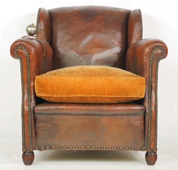 Vintage leather club chair with a plush amber seat cushion, studded trim, and a small ashtray on a strap at the left arm.