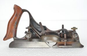 Side view of an antique plow plane with a dark wood handle, steel body, and visible depth adjustment and sliding skates, showing surface wear and a chipped handle.