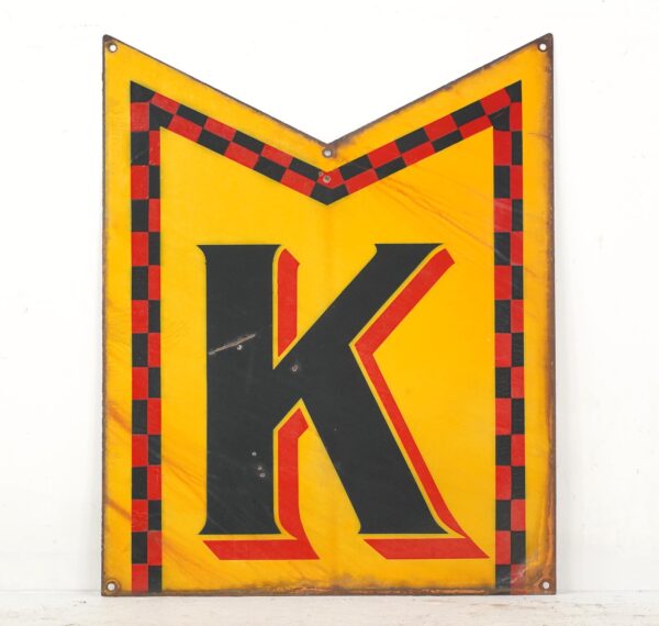 Pennant-shaped enamel sign with a large K in black, red, and yellow, showing heavy wear and warping, mounted with visible corner holes.