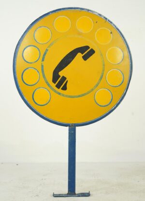 Vintage public telephone sign with a round yellow face, blue rotary dial rings, and a black handset icon on a blue steel post.