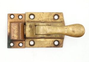 Antique brass transom window latch with a spring-loaded lever and mounting holes, shown in a close-up view with visible surface wear.