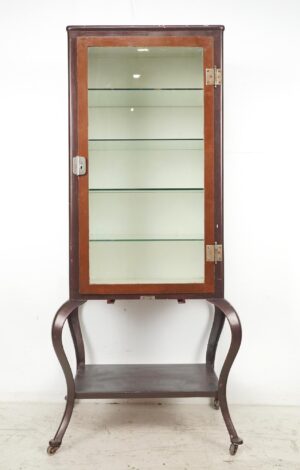 Tall vintage medical display cabinet with a glass door, four glass shelves, and curved legs on casters, shown in a front view with visible wear on the brown exterior finish.
