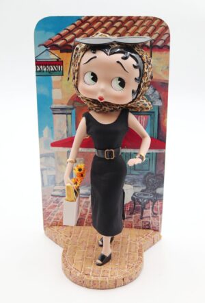 Porcelain collector doll in a black dress and leopard headscarf, holding a small shopping bag, posed on a cardboard stand with an Italian café scene backdrop.