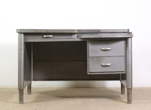 Gray steel tanker desk with an asymmetric top, three drawers on one side, and a lower side tray, shown front on with visible wear and rust spots.