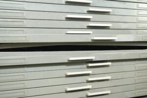 Gray steel flat filing cabinet with multiple drawers and several protruding pull handles, shown in close-up with one drawer slightly open.