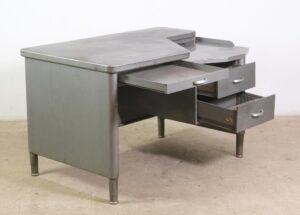 Gray sheet steel tanker desk with an asymmetric top, three drawers, and a lower tray section, shown at an angle with visible surface wear.