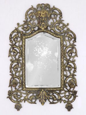 Rococo Revival wall mirror in antique brass with an ornate cast frame, Bacchus face at the top, and a beveled mirror plate showing age spots.