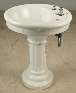 Vintage Art Deco white ceramic pedestal sink with a fluted column base, showing two faucet openings, remaining chrome hardware, and visible surface wear.